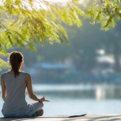 mulher de costas, sentada em frente a um lago, meditando.
